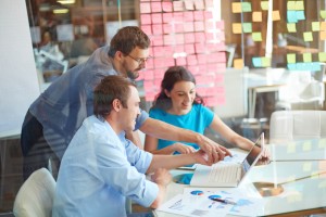 Group of three successful business partners in casual looking at laptop screen while two young men pointing at it at meeting in office