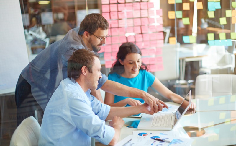 Group of three successful business partners in casual looking at laptop screen while two young men pointing at it at meeting in office